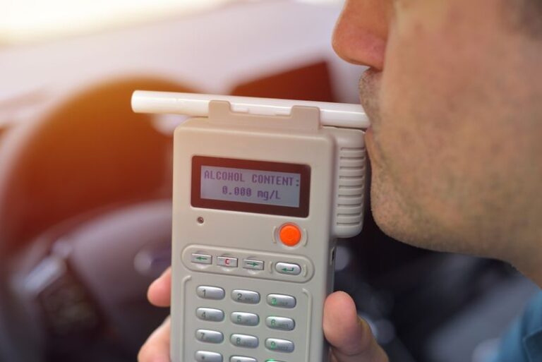Image is of a male driver blowing into a breathalyzer machine while in his car.
